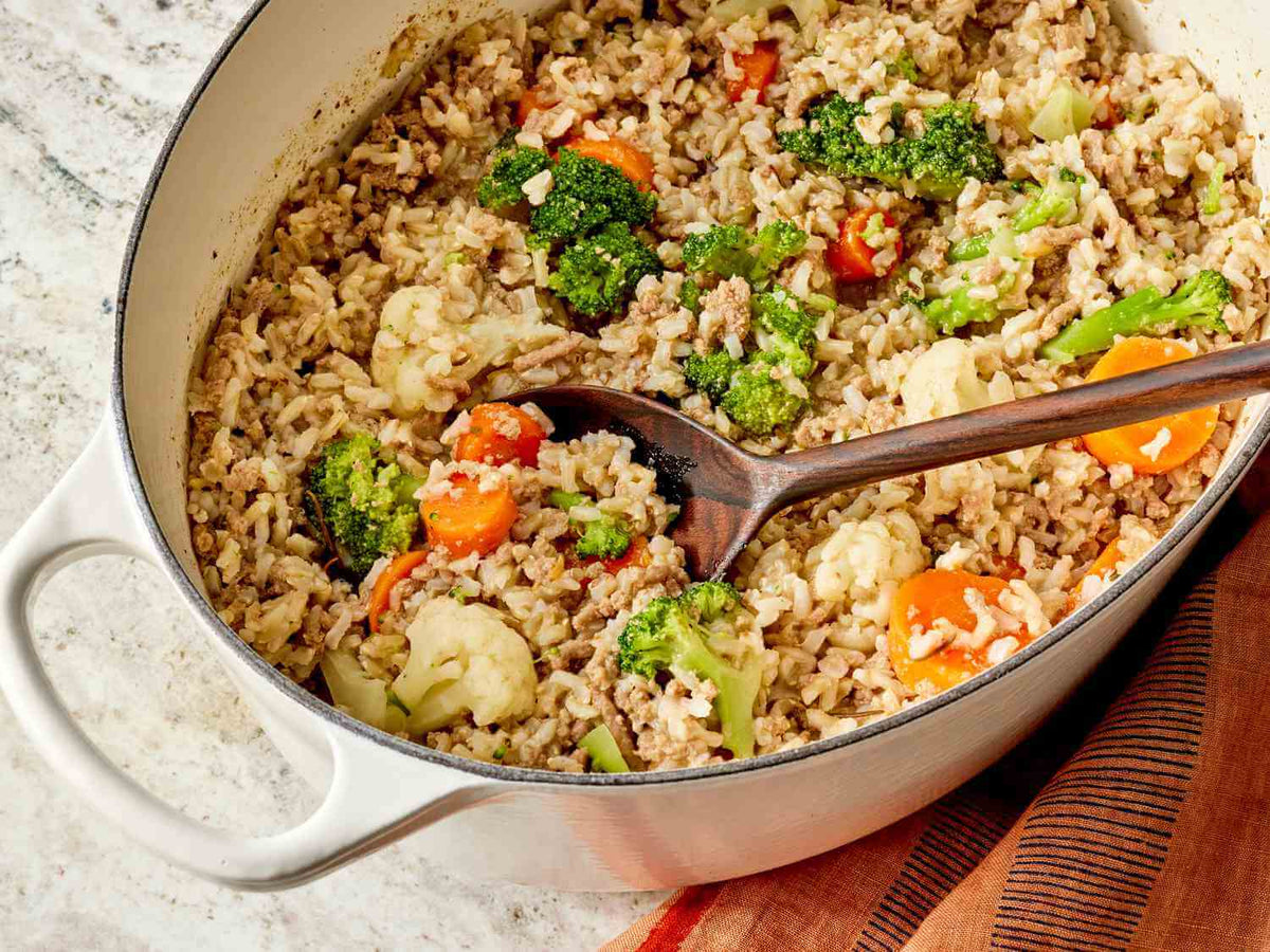 A large white Dutch oven filled with batch-cooked homemade dog food made with ground meat, brown rice, broccoli, carrots, and cauliflower, representing a cost-effective way to cook for your dog at home.