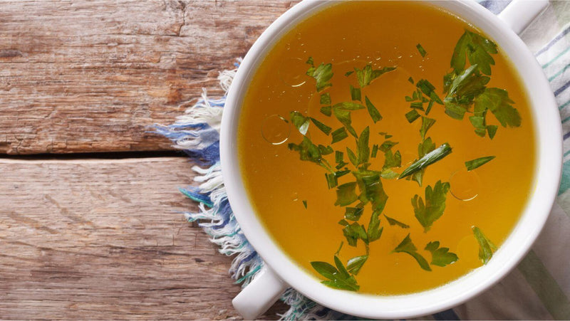 Golden bone broth with fresh herbs in a white ceramic bowl on a rustic wooden surface