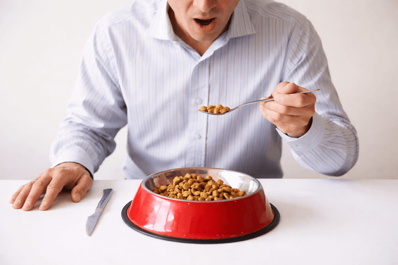 A person in a striped shirt uses a fork and knife to cut kibble in a red pet food bowl on a white background, adding humor to the scene.