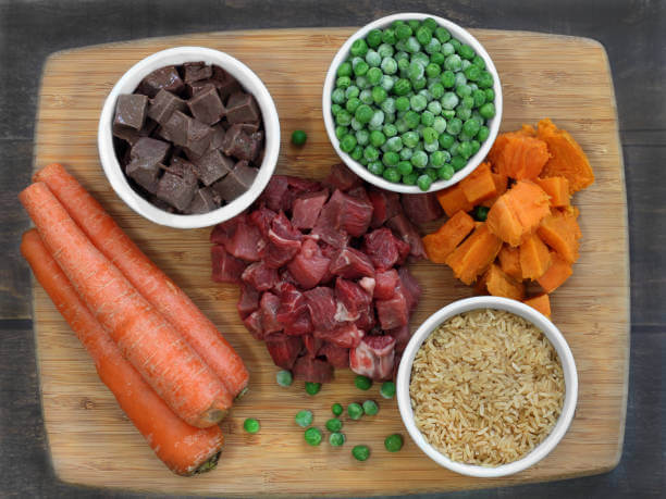 A wooden board holds fresh ingredients: carrots, diced beef, sweet potatoes, peas, rice, and liver cubes. The arrangement suggests a colorful, nutritious meal prep.
