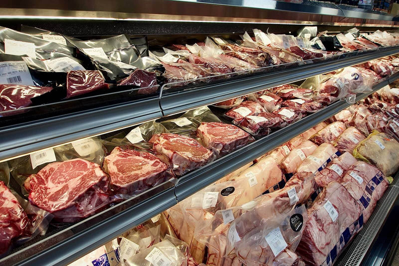 A well-stocked butcher shop refrigerator display filled with vacuum-sealed cuts of beef and other proteins, representing an affordable source for buying dog food proteins in bulk from local suppliers.