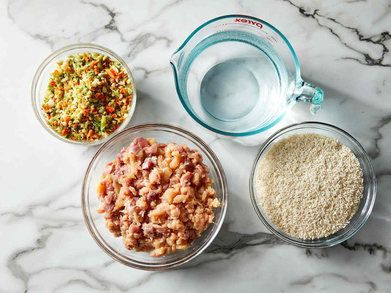 Raw ground chicken, mixed chopped vegetables, brown rice, and water in glass bowls on a marble surface, representing fresh whole-food ingredients used in gently cooked dog food recipes