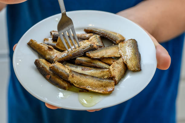 A person holds a white plate of grilled sardines, with a fork placed on top. The fish glisten with oil, against a blurred blue background, creating a fresh and savory scene.