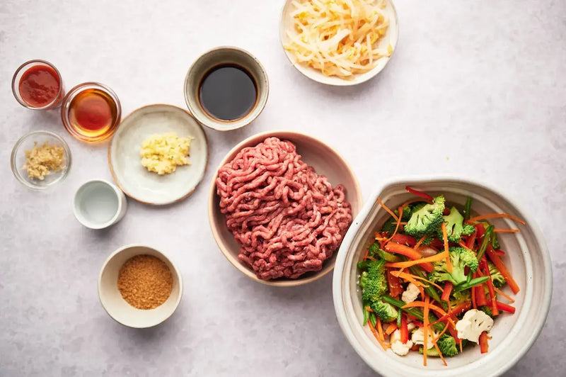 Flat lay of fresh dog food ingredients including ground meat, broccoli, carrots, red peppers, and various liquid seasonings in bowls on a light grey surface, representing nutrient-dense whole-food ingredients used in fresh dog food preparation. 