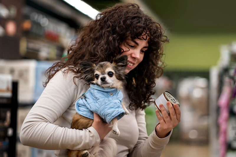Woman holding a small Chihuahua while reading the label on a can of dog food in a pet store, representing informed dog food purchasing decisions