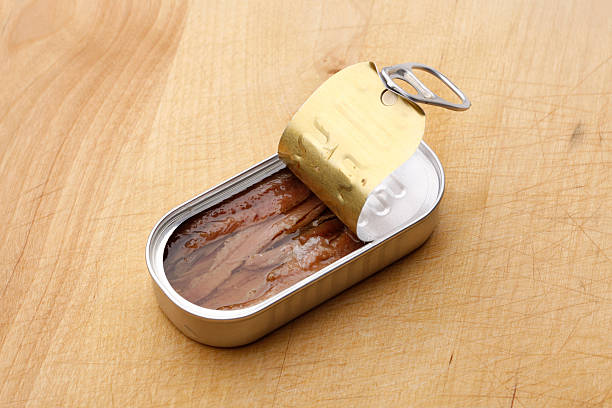 Open tin of anchovies on wooden surface. The metal lid is partially peeled back, revealing neatly packed fish fillets in oil. Simple and appetizing.