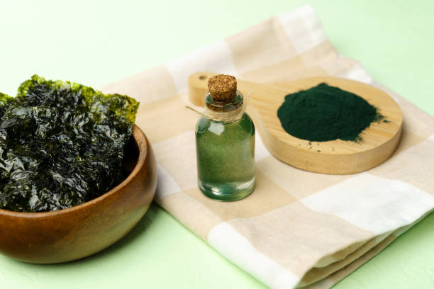 A wooden bowl of dried seaweed, a small corked glass bottle, and a wooden spoon with green powder sit on a checkered cloth, evoking a natural, earthy feel.